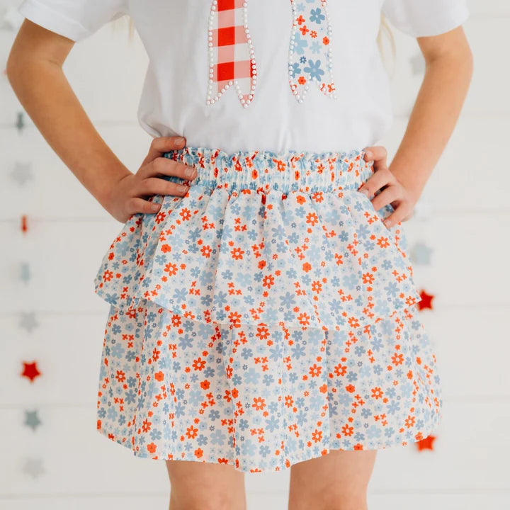 Model wearing Sweet Wink patriotic floral skort with a patchwork bow patch short-sleeve t-shirt, showcasing a vibrant, casual summer look.