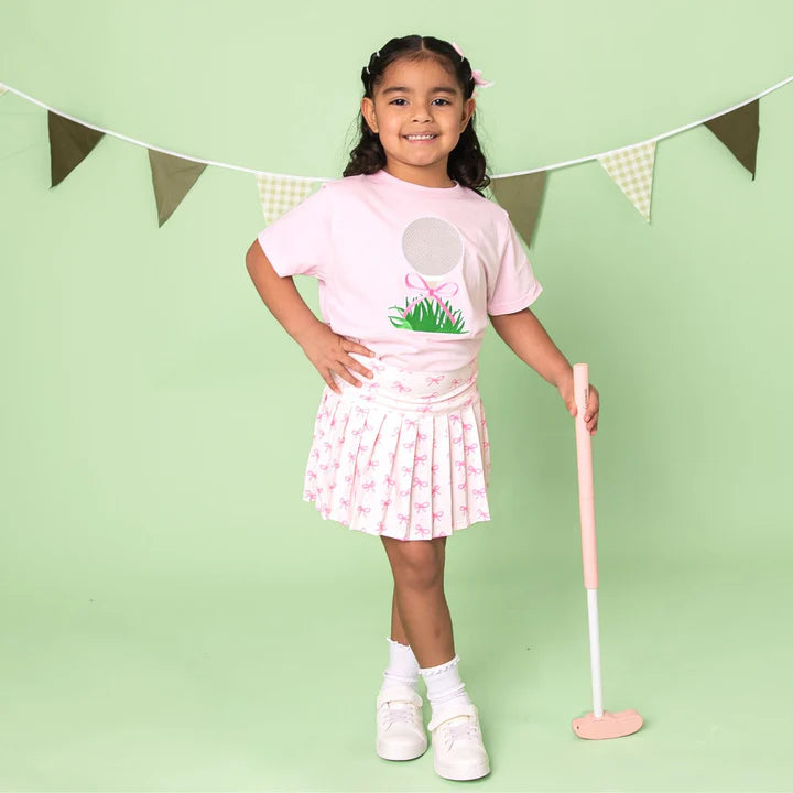 Model wearing a pink short sleeve T-shirt with a golf tee embroidered lace bow patch by Sweet Wink, styled with athletic skort.