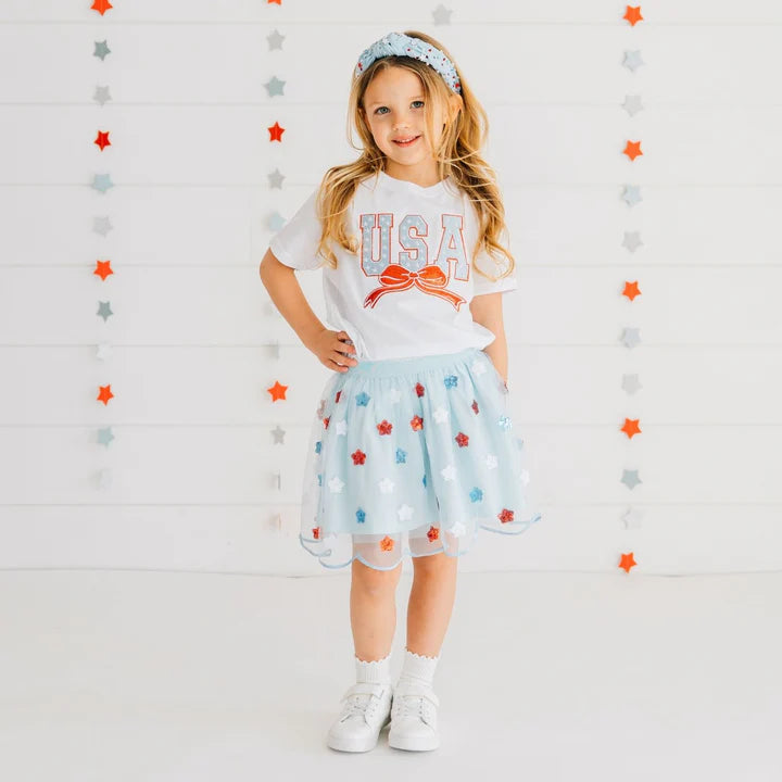 Model wearing a white Sweet Wink USA Stars & Bow Patch short sleeve T-shirt with sequin stars, gemstone headband, and tutu.