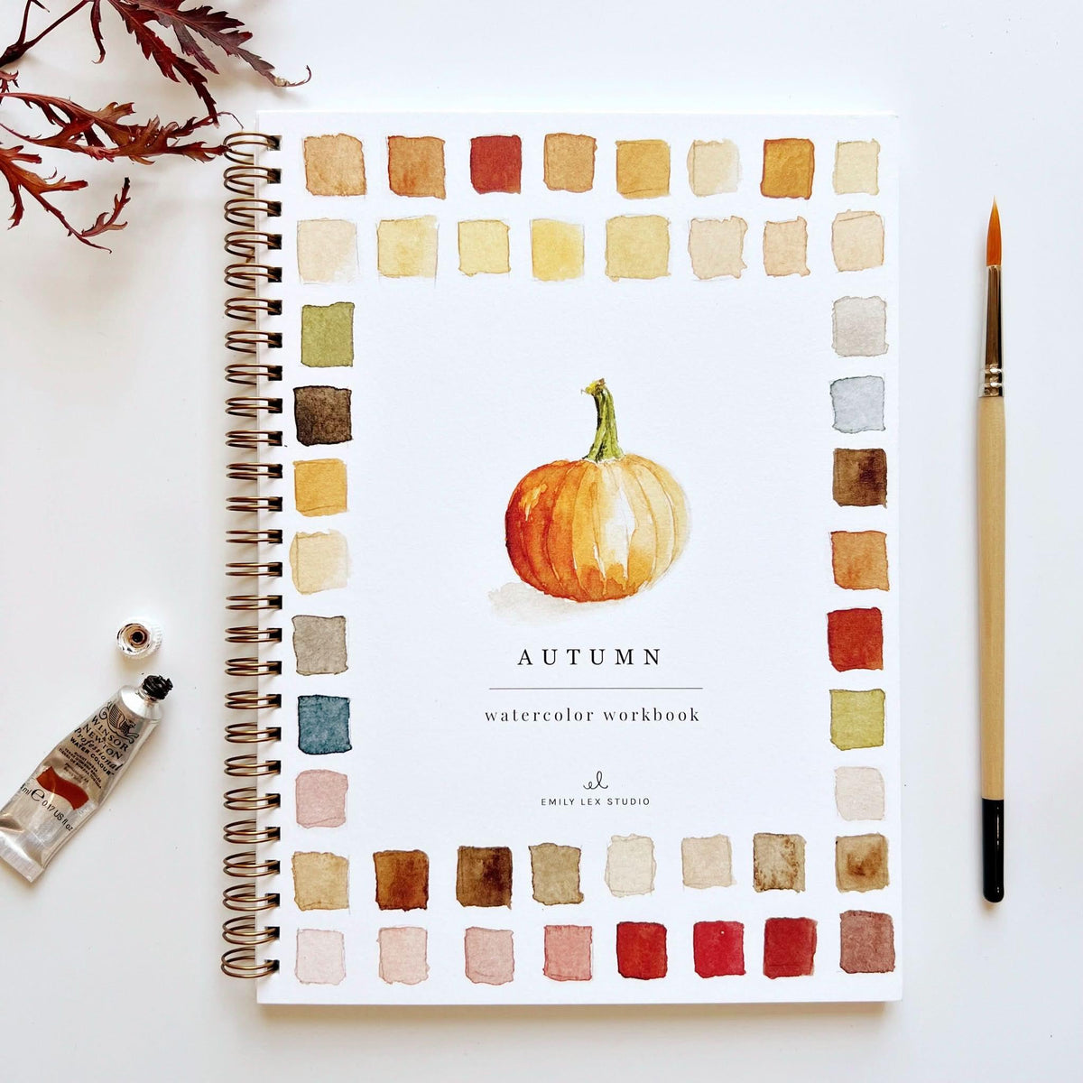 Watercolor workbook by Emily Lex Studio showing colorful pages and watercolor art supplies on a wooden table.